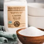 A close up of a bowl filled with India Tree Superfine Caster Sugar, positioned in front of its 1 lb bag, highlighting this premium caster sugar.