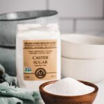 A bowl filled with India Tree Superfine Caster Sugar, positioned in front of its 1 lb bag, highlighting this premium caster sugar.